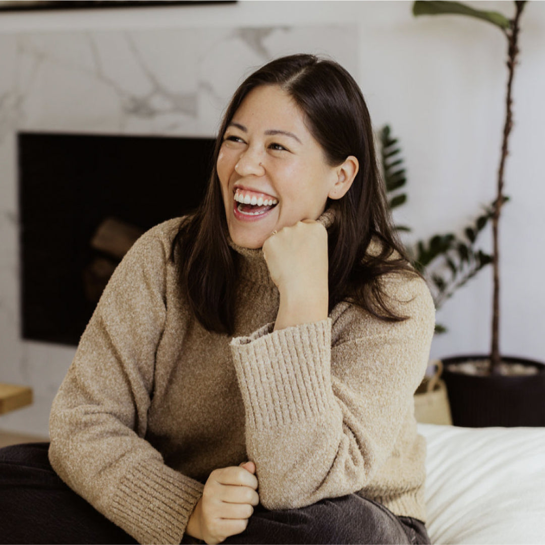 A woman with long dark hair, wearing a beige sweater and dark pants, sits and laughs. She rests her chin on her fist. Theres a fireplace and a potted plant in the background.